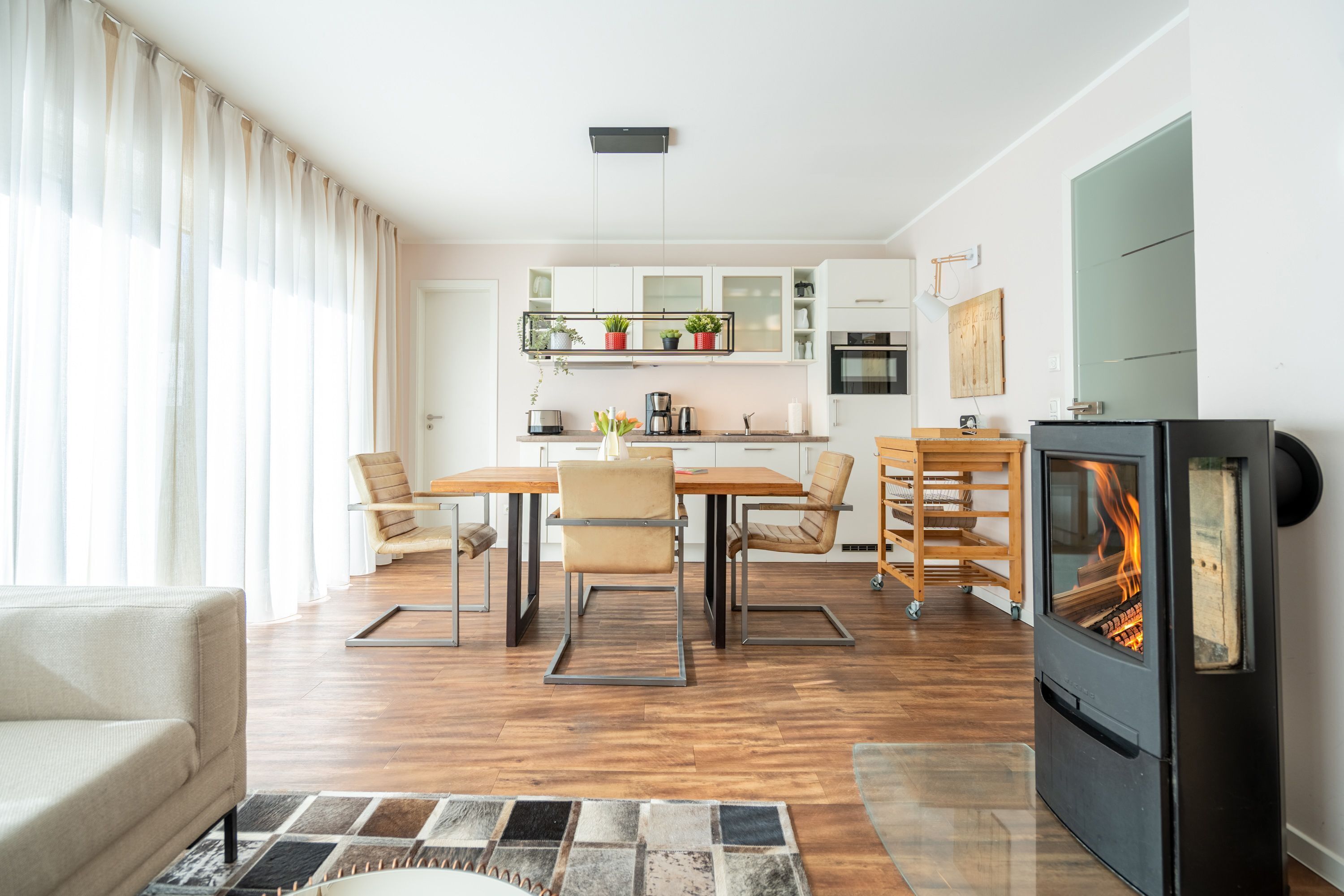 A modern living room with a sofa, a dining area with chairs and a table, a kitchen with cabinets and appliances, and a wood-burning stove.