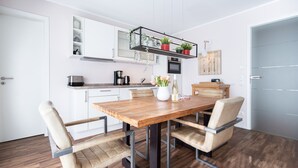 A modern kitchen with white cabinets, a wooden dining table, and four chairs. There is a coffee machine, a microwave, and a shelf with potted plants. - Quartier Ahlbeck, app. 04 (Heringsdorf)