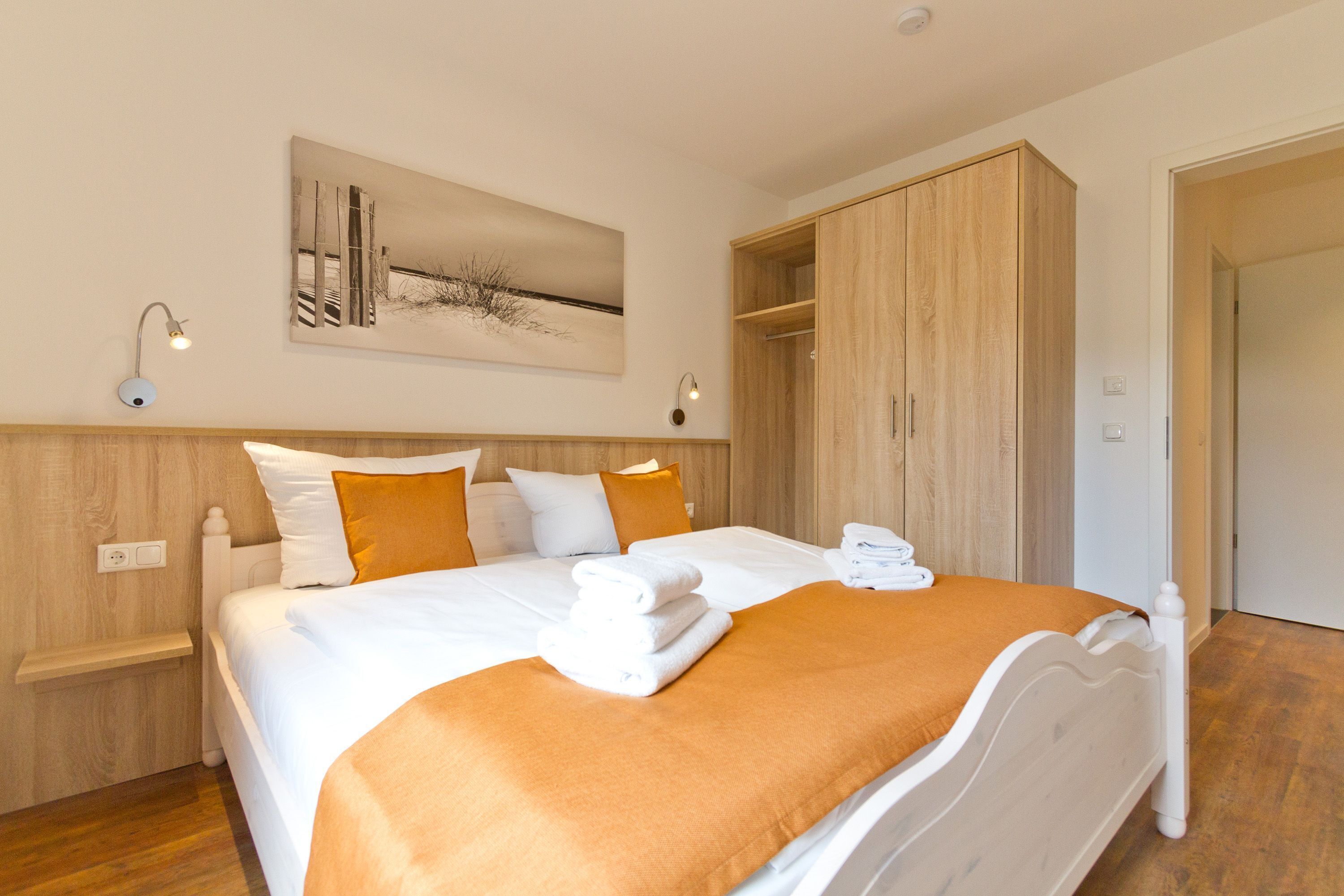 Een net opgemaakt bed met witte lakens en een oranje plaid, een houten kledingkast en een wandlamp.