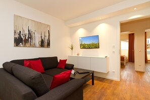 A modern living room with a grey sofa, red cushions, a coffee table, a flat-screen TV, and a cityscape painting. - Quartier Ahlbeck, app. 02 (Heringsdorf)