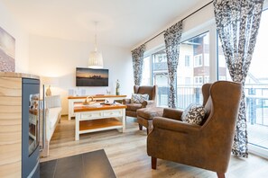 Una sala de estar moderna con un sofá, un sillón y una mesa de centro. Hay un ventanal con cortinas, un cuadro en la pared y una lámpara colgante.