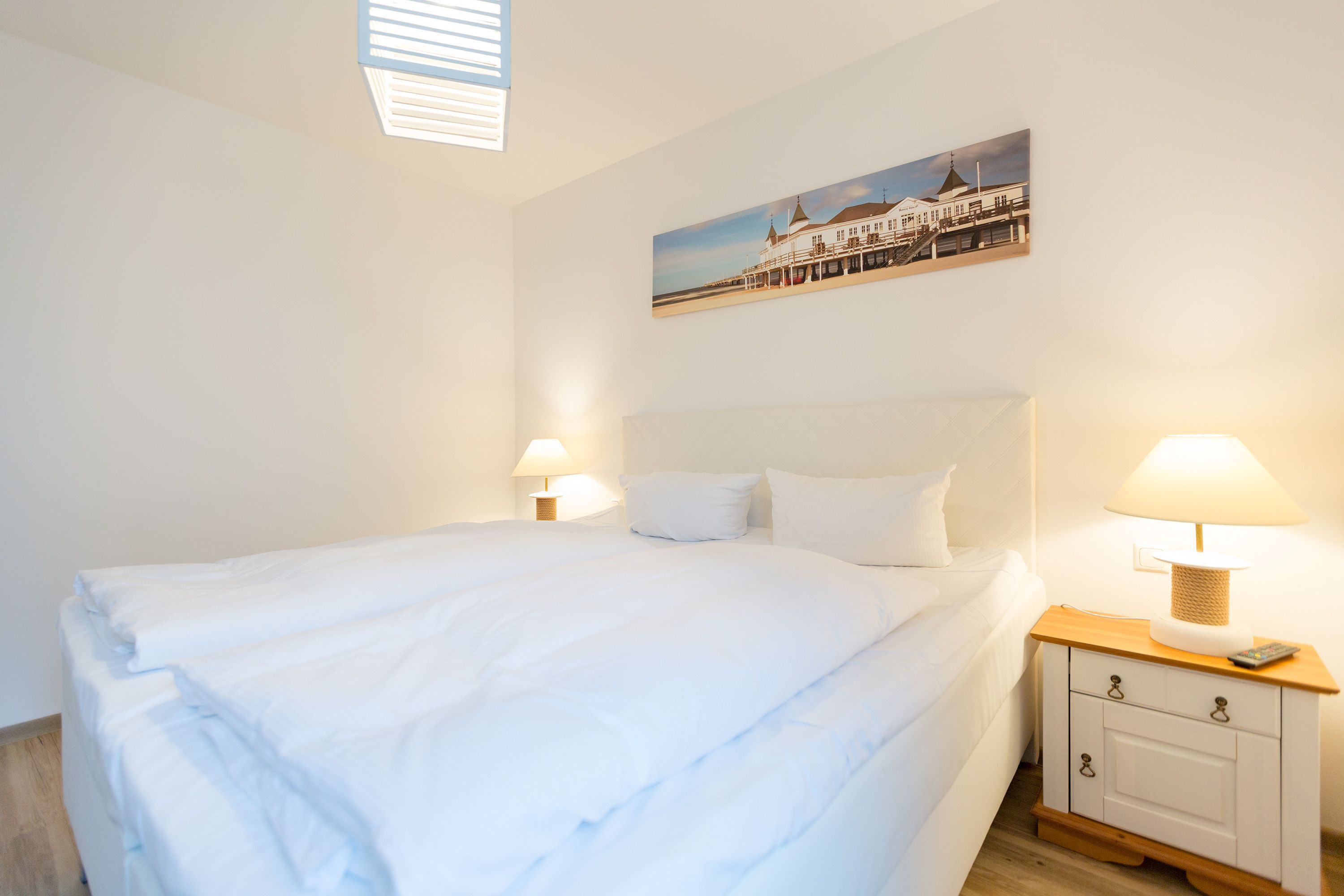 A neatly made bed with white linens, a bedside table with a lamp, and a painting of a building on the wall.