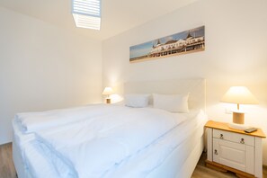 Una cama bien hecha con ropa blanca, una mesita de noche con una lámpara y un cuadro de un edificio en la pared.