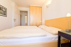 A hotel room with two beds, a wooden wardrobe, a bedside table with a lamp, and a framed picture on the wall.