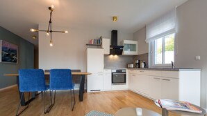 A modern kitchen with white cabinets, a built-in oven, and a window with white blinds. There is a dining area with blue chairs and a small table.
