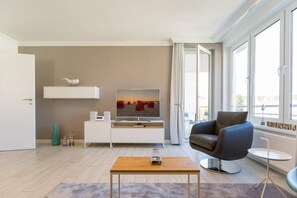 A modern living room with a flat-screen TV, a sofa, a coffee table, and large windows offering a city view.
