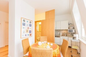 A compact kitchen with modern appliances, a dining area with a round table and wicker chairs, and a wall with framed certificates.
