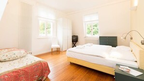 A bedroom with a bed, a chair, a dresser, a TV, and two windows. - Villa Astrid, T3 Münster (Heringsdorf)