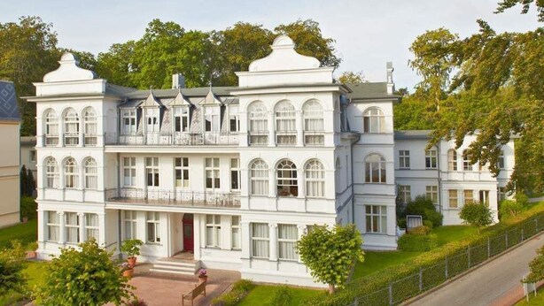 A large, white, two-story building with multiple windows and balconies, surrounded by greenery and a paved driveway. - Villa Astrid, T3 Münster (Heringsdorf)