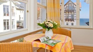A room with a table set for two, a wicker chair, a window with a view of a traditional building, and a vase with flowers. - Villa Astrid, T3 Münster (Heringsdorf)