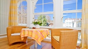 A room with two wicker chairs, a table with a checkered tablecloth, and a window with a view of buildings and trees. - Villa Astrid, T3 Münster (Heringsdorf)