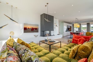 Un salon moderne avec un canapé vert, une table basse et une cheminée. On y trouve également un coin repas avec des chaises rouges et, au fond, une cuisine.