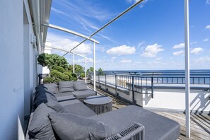 Ein Balkon mit einer Couch und einem runden Tisch, mit Blick auf einen Strand und das Meer.