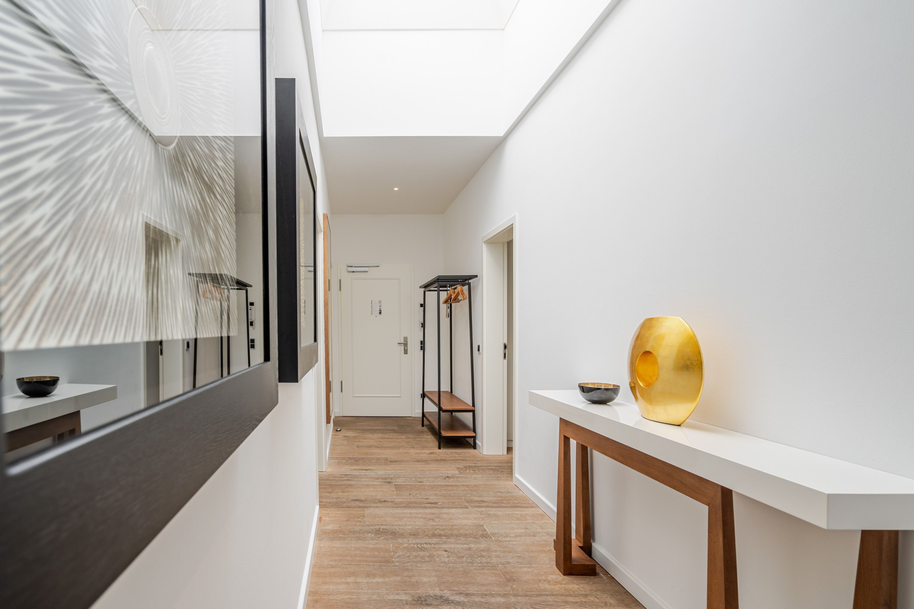 Een moderne hal met houten vloer, een witte muur en een dakraam in het plafond. Er staat een witte console tafel met een gouden vaas en een zwarte kom.