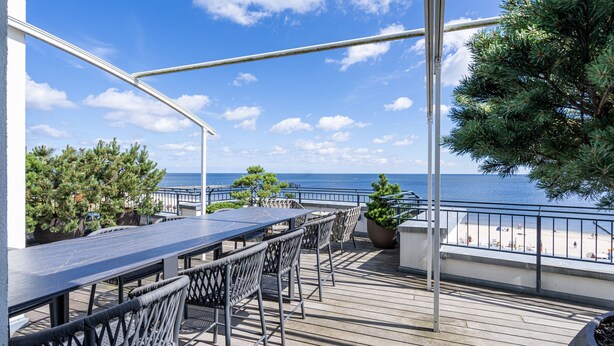 Eine Terrasse im Freien mit einem langen Tisch und Stühlen, mit Blick auf einen Strand und das Meer.