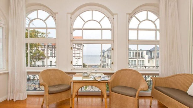 A room with three arched windows, wicker furniture, and a view of buildings and water. - Villa Astrid, T1 Cologne (Heringsdorf)