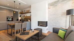 Ein modernes Wohnzimmer mit einer Couch, einem Couchtisch und einem Kamin. Es gibt einen Essbereich mit Tisch und Stühlen. Im Hintergrund ist eine Küche mit modernen Geräten zu sehen.