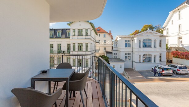Ein Balkon mit Tisch und Stühlen, der auf eine Straße mit parkenden Autos und historischen Gebäuden blickt.