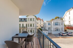 Uma varanda com mesa e cadeiras, com vista para uma rua com carros estacionados e edifícios históricos.