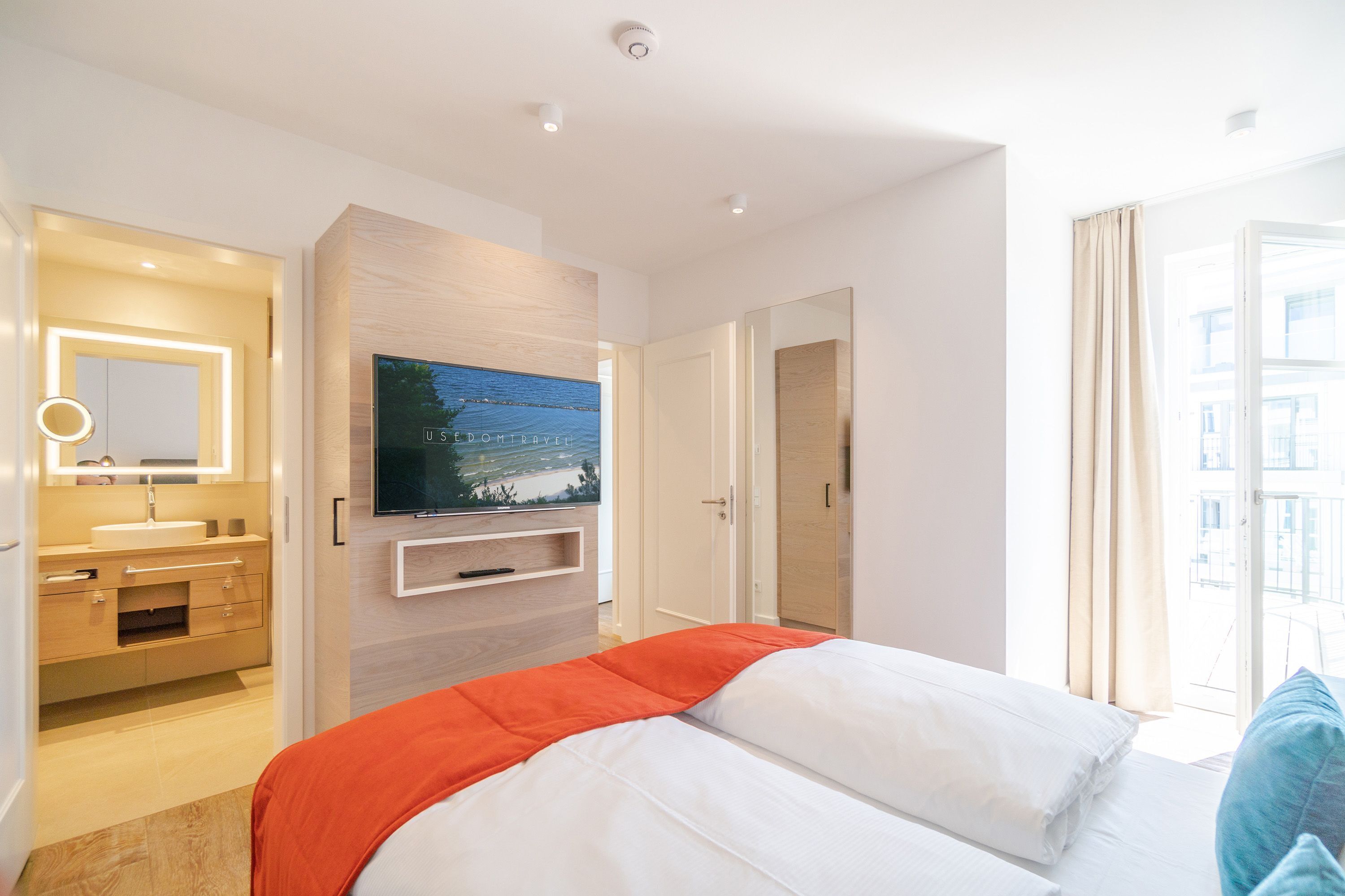 A modern hotel room with a bed, a TV mounted on the wall, a bathroom with a vanity, and a view of the city through the window.
