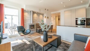 A modern living room with a sofa, a coffee table, and a TV. There is a fireplace and a dining area in the background. - Beachhouse Bansin, App. 21 (Heringsdorf)