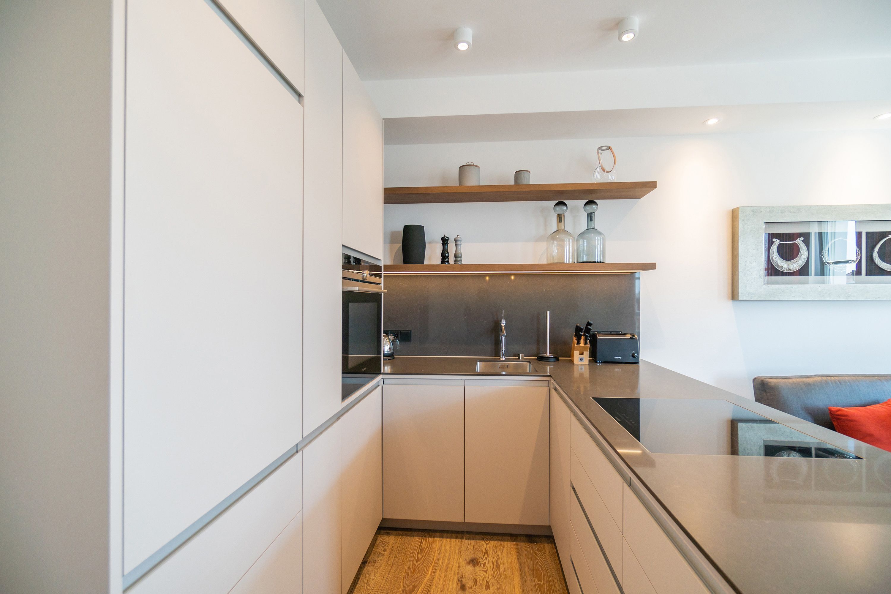 Eine moderne Küche mit weißen Schränken, einer Holz Arbeitsplatte und einem eingebauten Backofen. Über der Spüle befinden sich Regale mit Dekoration und ein Gemälde an der Wand.