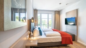 A modern hotel room with a large bed, a TV mounted on the wall, a window with curtains, and a wooden floor. - Beachhouse Bansin, App. 19 (Heringsdorf)