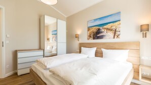 Een slaapkamer met een groot bed, een houten hoofdbord, een nachtkastje, een spiegel en een schilderij van een zandstrand.