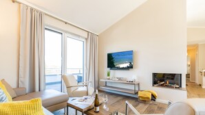 Een moderne woonkamer met een bank, fauteuil, salontafel, televisie, open haard en uitzicht op een balkon.