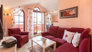 Een gezellige woonkamer met een rode bank, een witte salontafel en een rode fauteuil. Er is een groot raam met gordijnen, een schilderij aan de muur en een lamp aan de linkerkant.