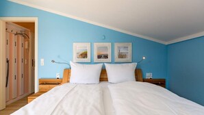 Een tweepersoonsbed met wit beddengoed in een kamer met blauwe wanden. Daarboven hangen drie ingekaderde foto’s.