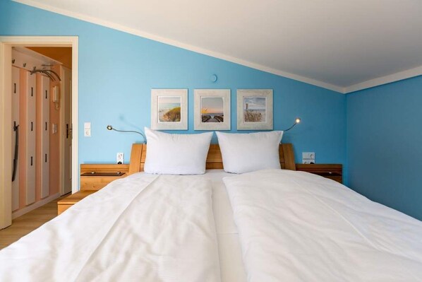 Uma cama de casal com roupa de cama branca num quarto com paredes azuis. Há três quadros na parede acima da cama.