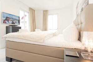 Ein modernes Hotelzimmer mit einem großen Bett, einem Flachbildfernseher, einem Nachttisch mit Lampe und Blick auf die Stadt durch das Fenster.