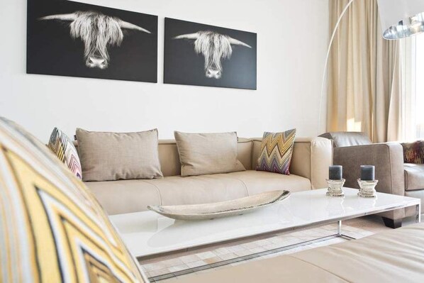 Ein modernes Wohnzimmer mit einem beigen Sofa, zwei schwarz-weißen Kunstwerken, einem weißen Couchtisch mit Kerzen und einer Stehlampe.