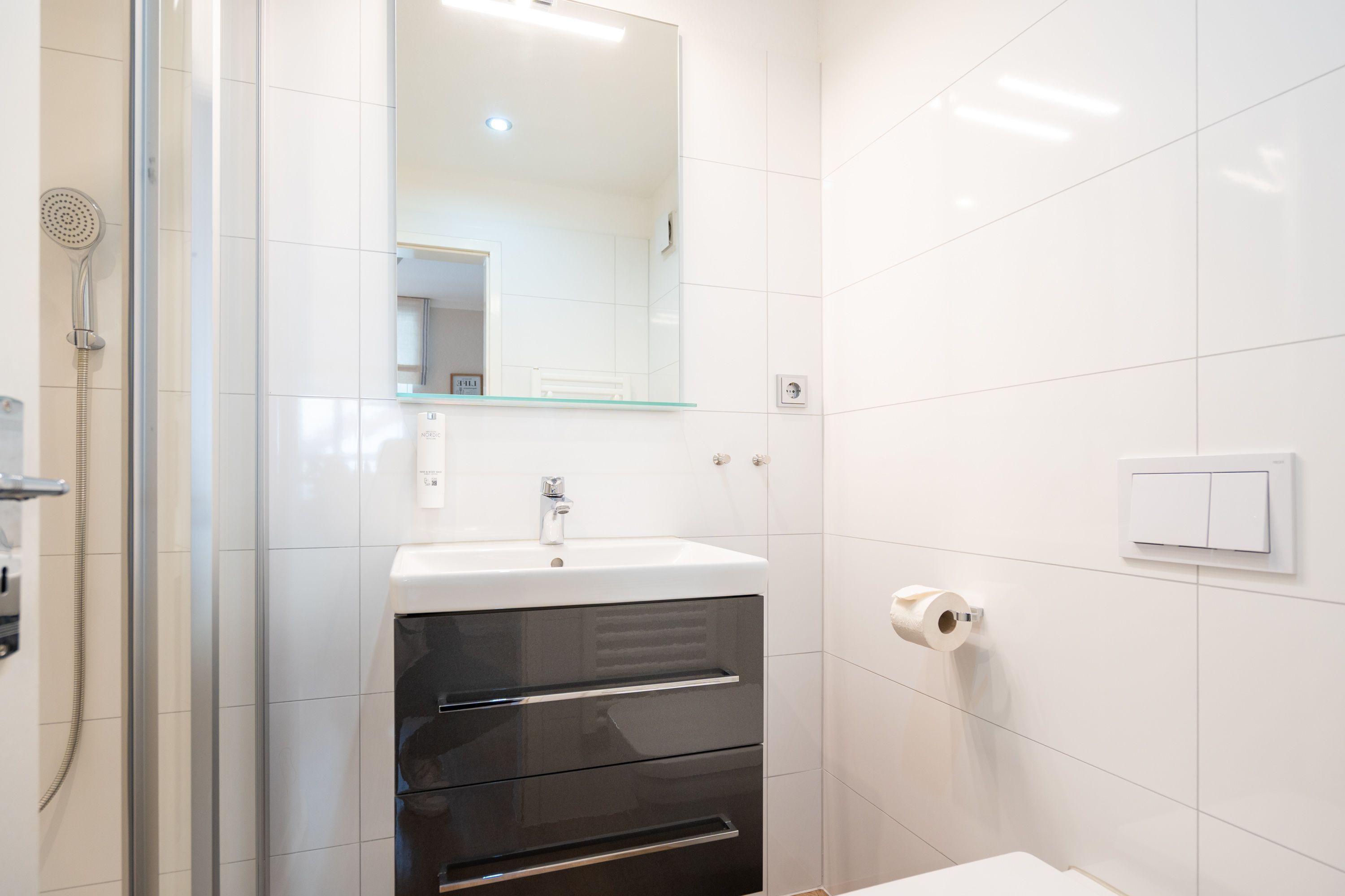 Ein modernes Badezimmer mit einer Duschkabine aus Glas, einem weißen Waschbecken mit Spiegel darüber und einem Toilettenpapierhalter an der Wand.