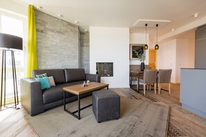 Un salon moderne avec un canapé gris, une table basse en bois et un pouf gris. On aperçoit à l’arrière-plan un coin repas avec une table et des chaises.