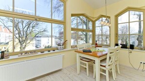 Ein Essbereich mit Tisch und Stühlen, große Fenster mit Blick auf Gebäude und Bäume sowie eine hängende Leuchte.
