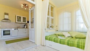 Ein Schlafzimmer mit einem Bett, einem Fenster mit Vorhängen, einem Nachttisch und Blick zur Küche.
