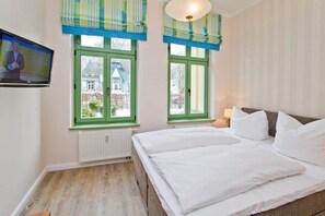 Ein Schlafzimmer mit einem Bett, zwei Fenstern mit grünen Rahmen, einem Heizkörper und einem Fernseher an der Wand.