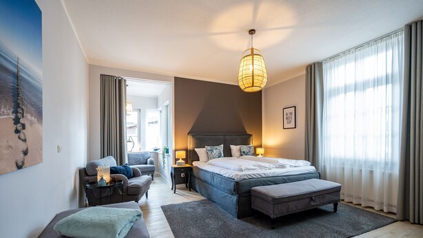 Una habitación de hotel moderna con una cama grande, una zona de estar con sofá y sillón, una mesita con lámpara, y vistas al exterior a través de amplios ventanales.