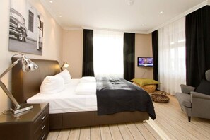 A modern hotel room with a large bed, a desk with a lamp, a television, and a comfortable armchair.