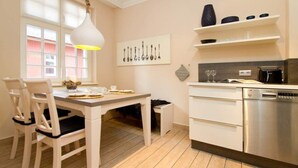 A modern kitchen with white cabinetry, a dining table with chairs, a pendant light, and a window with blinds.