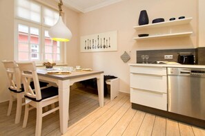A modern kitchen with white cabinetry, a dining table with chairs, a pendant light, and a window with blinds.