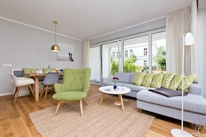 Un salon moderne comprenant un canapé, un fauteuil, une table à manger et de grandes fenêtres donnant sur des bâtiments et de la verdure.