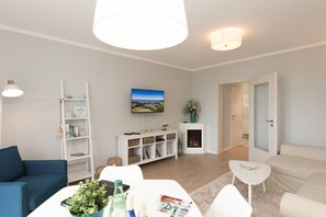 Uma sala de estar moderna com um sofá branco, uma poltrona azul, uma mesa de centro branca com revistas e uma planta, uma estante branca, uma lareira e uma televisão fixada na parede.