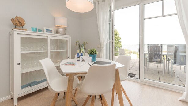 A dining area with a white table, four chairs, a cabinet with glass doors, and a view through a large window. - Haus am Schmollensee, app. 7h (Heringsdorf)