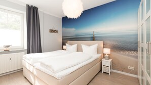 A bedroom with a large bed, a nightstand, a lamp, a window with curtains, and a beach mural on the wall. - Haus am Schmollensee, app. 7h (Heringsdorf)