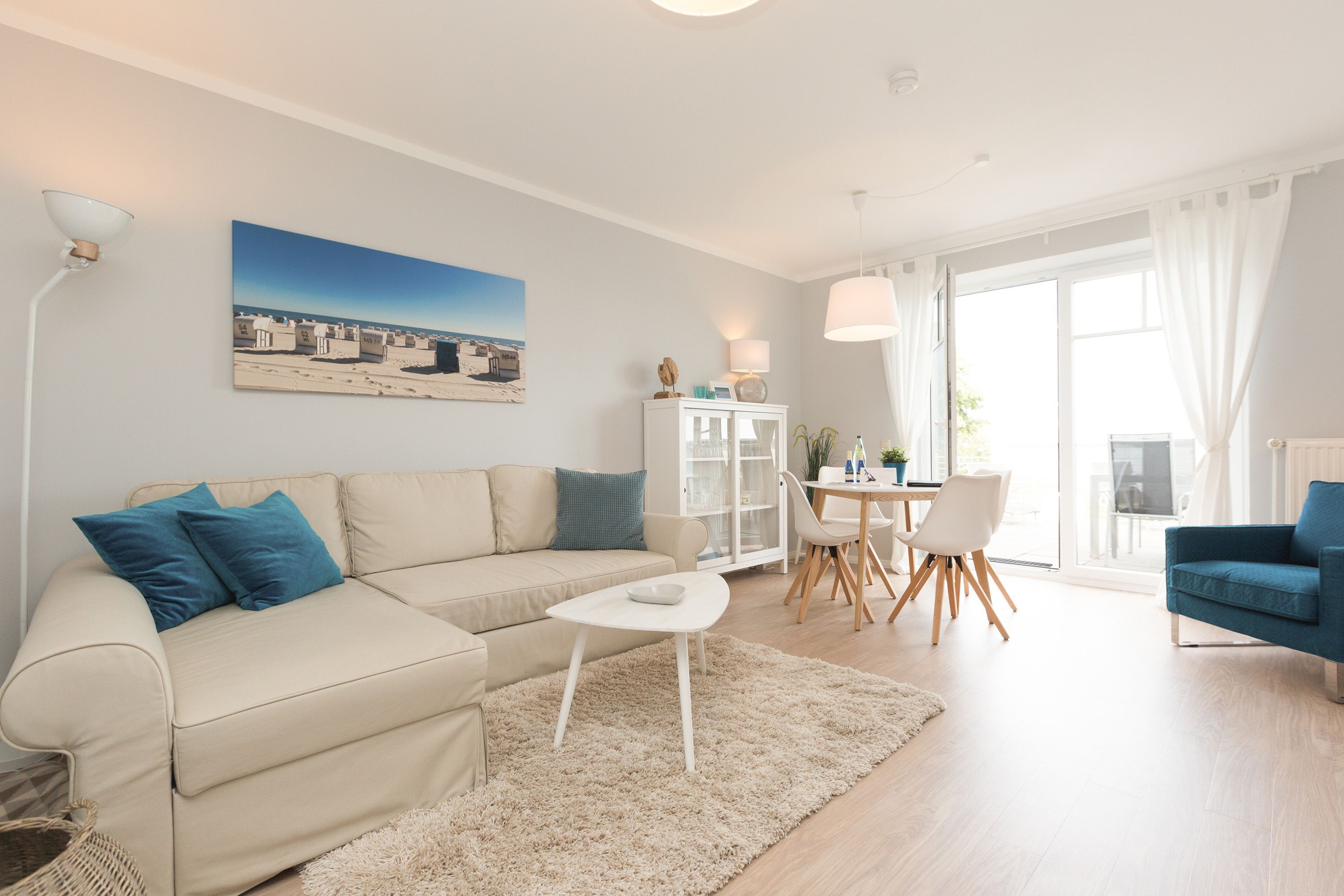 A living room with a beige sofa, a white coffee table, a dining table with chairs, and a large window with curtains.