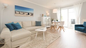 A living room with a beige sofa, a white coffee table, a dining table with chairs, and a large window with curtains. - Haus am Schmollensee, app. 7h (Heringsdorf)