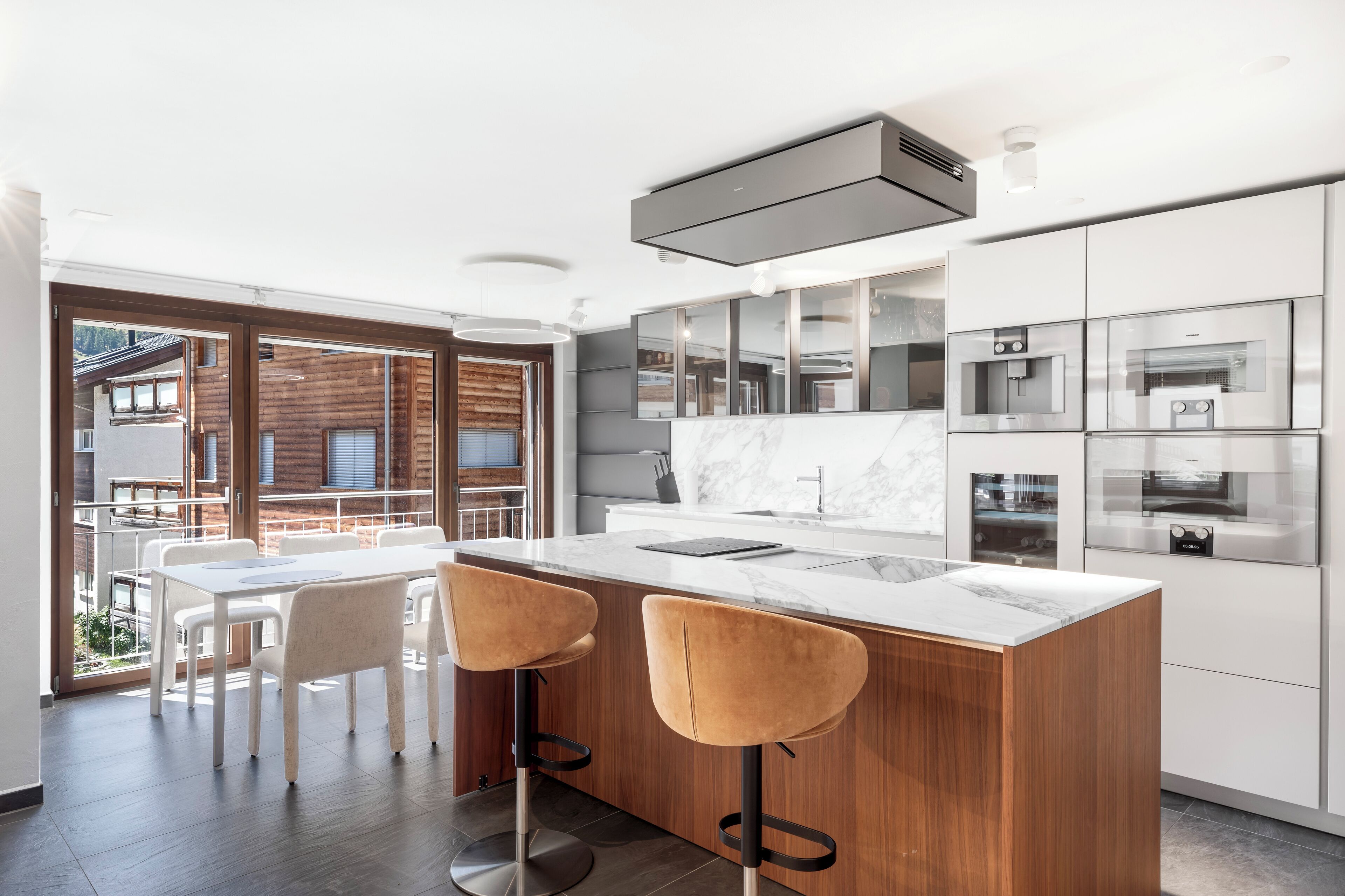 A modern kitchen with a marble countertop, wooden cabinets, and stainless steel appliances. There is a dining area with white chairs and a table.